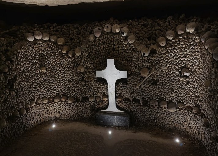 A pale stone cross set into a wall of stacked femurs and skulls in the Paris Catacombs