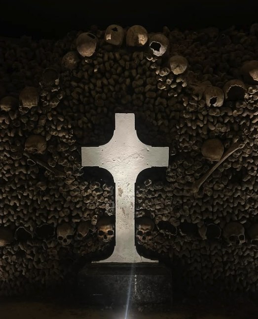 A pale cross standing against a dark wall of skulls and bones in the Paris Catacombs