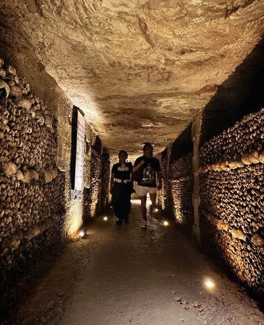 Two visitors walking a bone-lined gallery of the Paris Catacombs